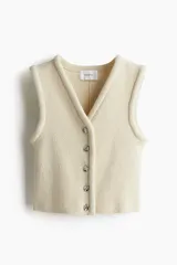 Chaleco corto de punto color crema, con escote en V, botones metálicos de diseño irregular y ribete tubular en escote, tapeta y sisas.