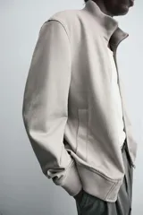 Cazadora color gris claro de corte regular, confeccionada en tejido con efecto ante. Tiene cuello alto, cierre frontal con cremallera, bolsillos laterales y puños y bajo en rib elástico.