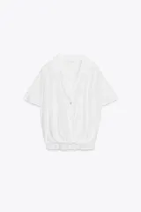 Camisa blanca de cuello solapa y escote pico, con mangas cortas y bajo asimétrico con nudo en el delantero. Cierre frontal con botones.