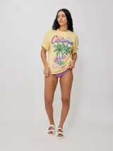 Remera color crudo de corte oversized con estampado de palmeras, flores y texto 'California' y 'Rusty Tribe'.