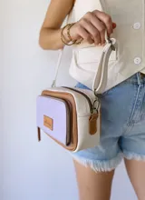 Bandolera rectangular de cuero color lila, con detalles en marrón y blanco. Tiene correa larga regulable, bolsillo frontal con cierre y etiqueta de cuero con logo de la marca.