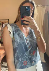 Chaleco sin mangas con escote en V y botones frontales, confeccionado en tela estampada con diseño tipo patchwork en tonos azules y grises, con detalles florales en rojo y amarillo. Presenta volados en los hombros.