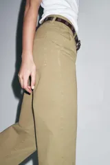 Pantalón cropped color beige de tiro alto y corte recto, confeccionado en algodón. Cuenta con bolsillos delanteros y traseros, trabillas en la cintura y cierre frontal con cremallera y botón.