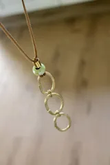 Collar con cordón de cuero marrón, cuenta con una cuenta de vidrio verde y tres dijes de círculos de bronce.