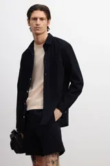 Camisa negra de sirsaca de algodón con cuello inglés, tapeta clásica, bolsillo superior de parche y canesú con pliegue de caja en la espalda. Mangas largas con tapeta de manga y botón, y cierre ajustable de botón en los puños. Bajo redondeado y corte holgado.