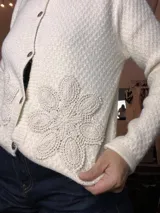 Cardigan de punto grueso color blanco roto, con botones oscuros y aplique floral de crochet en el lateral inferior derecho.