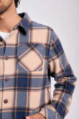 Sobrecamisa de tejido grueso a cuadros, con cuello camisero, botones frontales y bolsillo de parche en el pecho. El diseño combina tonos beige tostado, azul marino y marrón.