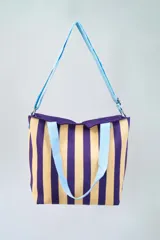 Bolso tipo tote con diseño a rayas verticales color beige y violeta. Tiene doble asa de mano celeste y correa larga al hombro también celeste.