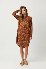 Vestido camisero corto color marrón con estampado abstracto en tonos naranja. Tiene cuello camisero, cierre frontal con botones y mangas largas.