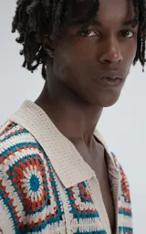 Camisa de manga corta, tejida a crochet con un patrón de cuadrados en colores beige, naranja y turquesa. Presenta cuello tipo polo y cierre frontal con botones.