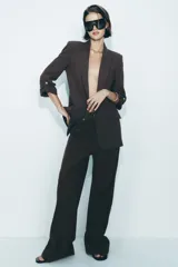 Blazer color marrón oscuro, abierta, con cuello solapa y hombreras. Manga tres cuartos con trabilla y botón dorado. Bolsillos delanteros con solapa.
