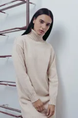 Vestido corto color beige de tejido de punto, con cuello alto tipo polera y mangas largas. Corte recto y terminaciones acanaladas en puños, cuello y ruedo.