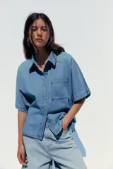 Camisa de denim celeste con cuello solapa, manga corta, bolsillo de parche en el pecho, bajo con aberturas laterales y cierre frontal con botones.
