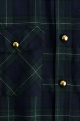 Camisa oversized de franela suave con estampado a cuadros en tonos azul marino, verde oscuro y beige. Presenta cuello, botonadura frontal con botones dorados, canesú en la espalda, hombros caídos, bolsillos superiores con solapa y botón dorado, y puños abotonados. El bajo es ligeramente redondeado.