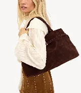 Cartera marrón de gamuza con doble asa corta, correa con hebilla decorativa y cierre superior.