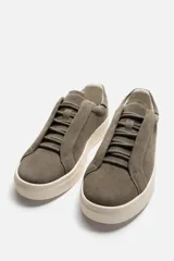 Zapatilla deportiva de hombre color gris topo, fabricada en piel con acabado serraje. Presenta cordonera elástica oculta y suela de goma de bajo volumen.