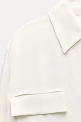 Camisa negra de satén con cuello solapa, manga larga, bolsillos con solapa en el delantero, bajo asimétrico y cierre frontal con botones.