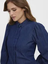 Camisa de denim color azul oscuro, de manga larga con hombros abullonados y puños ajustados con botón. Presenta cuello clásico y cierre frontal de botones.