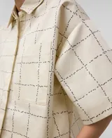 Camisa de algodón color crema con estampado de cuadros negros. Tiene cuello solapa, manga corta, bolsillo en el pecho y cierre frontal con botones ocultos.