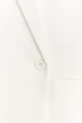 Blazer blanco de corte oversize, con cuello solapa, manga larga con hombreras, bolsillos de doble vivo en el delantero, aberturas en la espalda y cierre cruzado con botones.