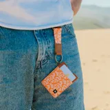 Llavero con mosquetón metálico color bronce, cinta de tela estampada en tonos coral y naranja, y pieza de cuero sintético marrón con logo grabado.