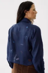 Camisa azul de lino con cuello clásico, manga larga y cierre frontal con botones. Presenta bordados de dados, cartas y el nombre de la marca.