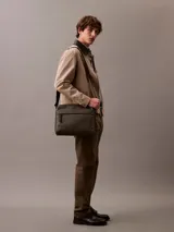 Bolso bandolera Calvin Klein de cuero sintético texturizado color verde oliva, con correa ajustable de tela marrón oscuro que lleva el logo de la marca repetido. Presenta un bolsillo frontal horizontal y el logo CK grabado en la parte inferior derecha.