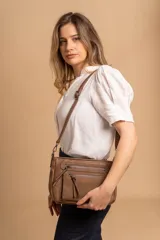 Bandolera de simil cuero color marrón topo, con textura ligeramente desgastada y detalles en nobuck. Presenta múltiples bolsillos frontales con cierres metálicos y tiradores de cuero. Correa ajustable para llevar al hombro o cruzada.