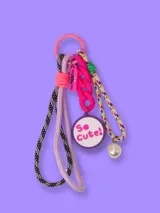Llavero con aro metálico rosado, cordones trenzados en blanco y negro y lila, cadena plástica rosada, dije redondo con la frase 'So Cute!' y una perla plástica.
