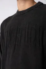 Sweater de punto grueso color gris oscuro con cuello redondo y mangas largas. Presenta un diseño de texto bordado en el frente y detalles de roturas en los puños y el ruedo.