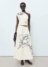 Top de algodón color crema, con cuello asimétrico, detalle de bordado floral en el hombro y frunces laterales. Cierre lateral con cremallera.