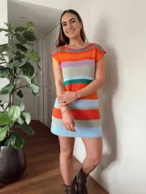 Vestido corto tejido con hilo de algodón a rayas horizontales en tonos celeste, azul, marrón, blanco, naranja y verde. Tiene cuello redondo y mangas cortas. El ruedo es desflecado.