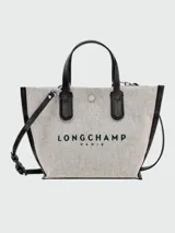 Bolso Longchamp modelo Essential Toile XS, color crudo con detalles en cuero negro. Tiene doble asa corta superior, correa larga ajustable y removible, cierre con broche a presión y logo estampado en el frente.