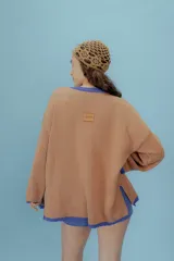 Buzo oversized de punto color tostado con diseño gráfico frontal en azul inspirado en una estampilla con una sirena y el texto 'Taormina'. Presenta tajos laterales, cuello redondo y terminaciones en contraste de color azul.