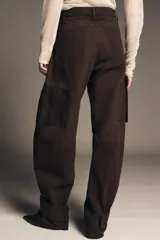 Pantalón cargo de algodón color marrón oscuro, tiro medio, con pliegues frontales, bolsillos delanteros ocultos y bolsillos de plastrón con solapa y botones a presión en las piernas. El bajo del pantalón tiene una trabilla con botón y se muestra doblado hacia afuera.