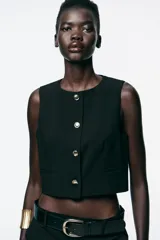 Chaleco corto negro con cuello redondo, sin mangas y cierre frontal con botones dorados.