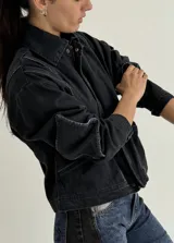 Campera de jean negra tipo bomber con cuello redondo y cierre frontal con botones metálicos. Presenta un parche rectangular con diseño gráfico en la espalda.