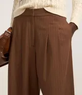 Pantalón de vestir marrón con finas rayas verticales blancas, de corte recto y tiro alto. Se ajusta a la cintura con cinturón de cuero marrón.