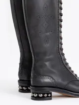 Bota de caña alta color negro, confeccionada en cuero vacuno, con cordones frontales marrones y cierre lateral. Presenta un diseño picado en la capellada y un taco de 4 cm con detalle de esferas metálicas.