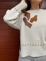Sweater color crema tejido en lana merino, con apliques de cuero marrón y tejido trenzado en el frente. Presenta costuras a la vista en el ruedo y los puños.