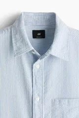 Camisa de hombre de corte holgado, confeccionada en tela de sirsaca a rayas verticales celestes y blancas. Tiene cuello inglés, tapeta clásica con botones, bolsillo de parche en el pecho, mangas largas con puños abotonados y bajo redondeado.