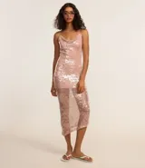 Vestido midi de malla con lentejuelas plateadas, escote recto y tirantes finos cruzados en la espalda. Presenta un forro corto que deja ver la transparencia de la malla con lentejuelas en la parte inferior, creando un bajo asimétrico.