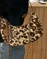 Cartera tipo shoulder bag con estampado animal print de vaca en tonos marrón y blanco, con correa de cuero y cadena metálica.