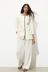 Chaqueta color crema de punto con cuello redondo y manga larga. Tiene bolsillos delanteros de plastrón, pespuntes combinados a contraste y cierre frontal con botones dorados.