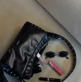 Bolso de hombro negro de cuero sintético, con forma rectangular y cierre superior. La correa es ancha y está decorada con tachas metálicas plateadas a lo largo de ambos bordes. El lateral del bolso también presenta tachas.