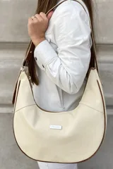 Cartera tipo hobo color crema con detalles en marrón. Tiene correa regulable para usar cruzada, forro interior con bolsillo y cierre, y bolsillo trasero con cierre.