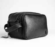 Neceser de viaje negro con textura símil cuero, cierre superior con doble cremallera y asa lateral.