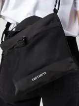 Bolso bandolera Carhartt WIP modelo Irwin, color negro. Cuenta con compartimento principal con cierre de cremallera, bolsillo lateral de malla, bolsillo frontal con cierre a presión, asa de mano y correa bandolera ajustable. Logo de la marca estampado en blanco en la parte inferior.