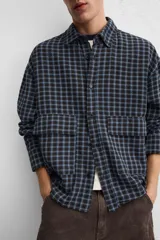 Camisa de hombre a cuadros azules y negros, de corte cuadrado, con cuello solapa, manga larga con puños abotonados, cierre frontal con botones y bolsillos de parche con solapa en el pecho.