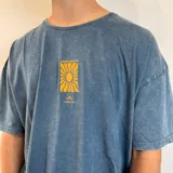 Remera azul con efecto gastado, corte oversize y estampa trasera con un sol amarillo dentro de un cuadrado negro y texto "This is living" y "Sunsets sessions".
